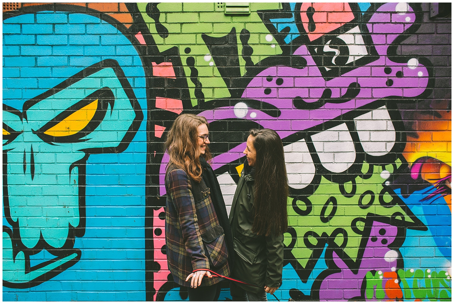 Same sex couple stand in front of a graffiti mural