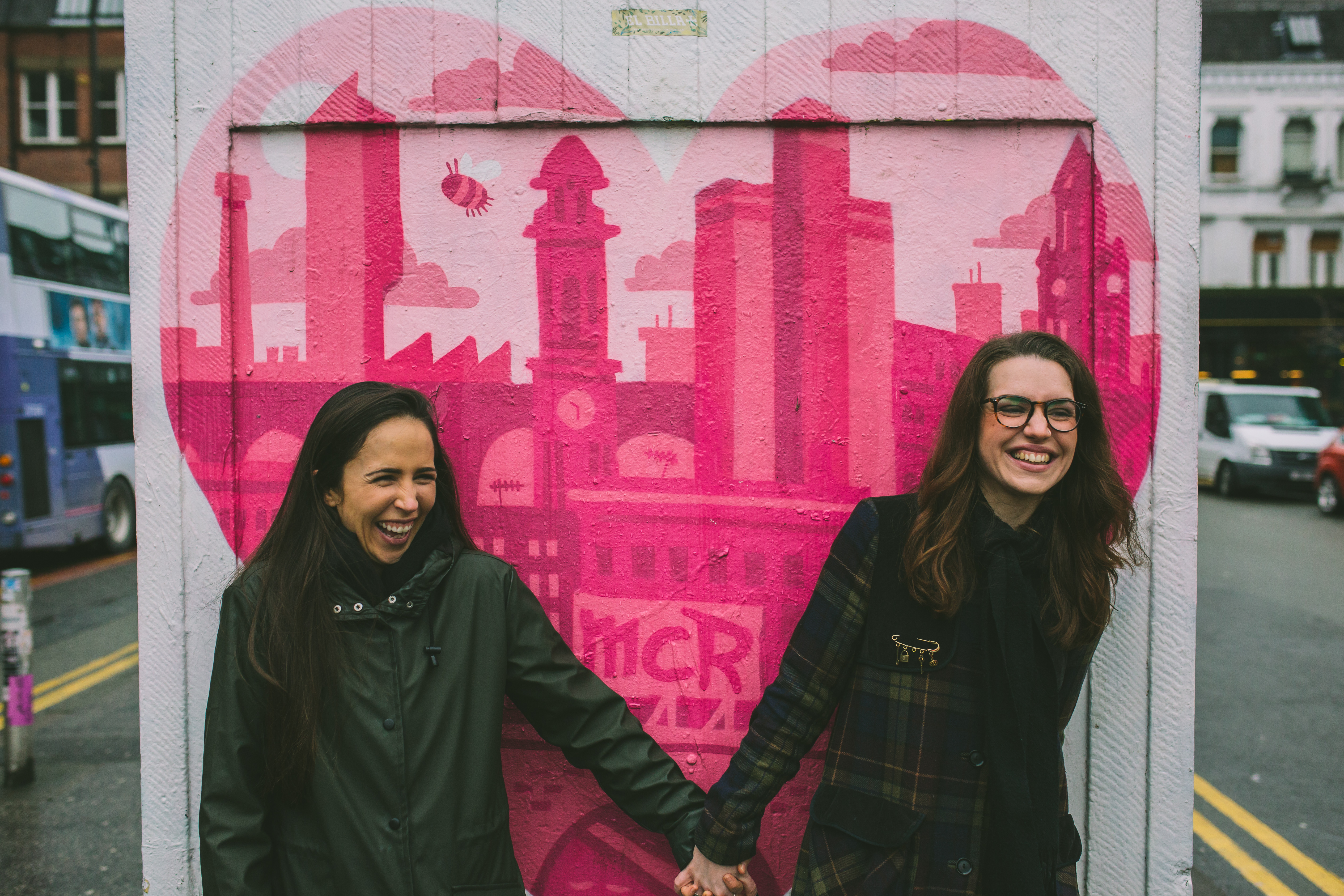 Female couple laugh holding hands in front of Manchester graffiti 