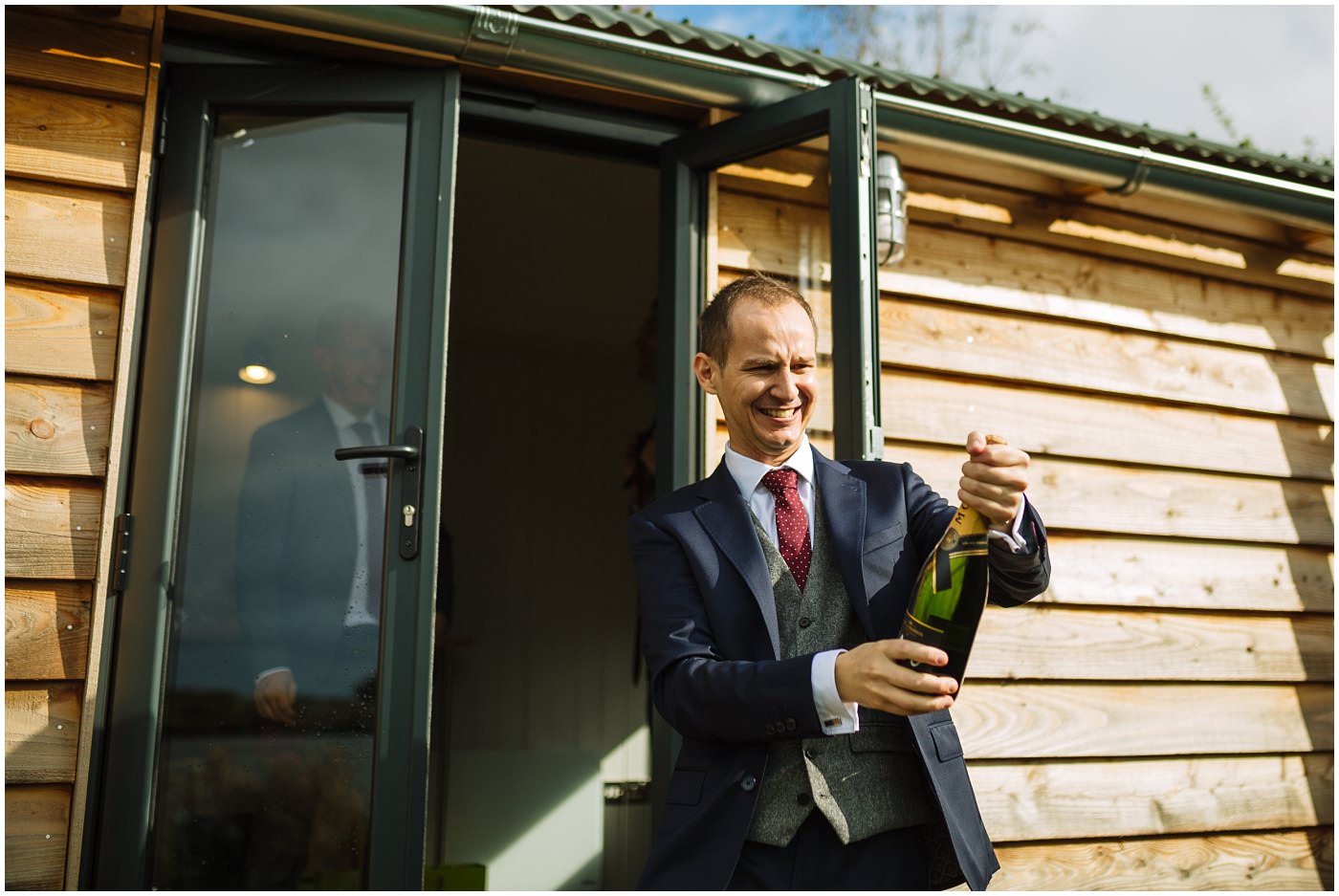 groom opens champagne in front of shepherds hut at sandhole oak barn