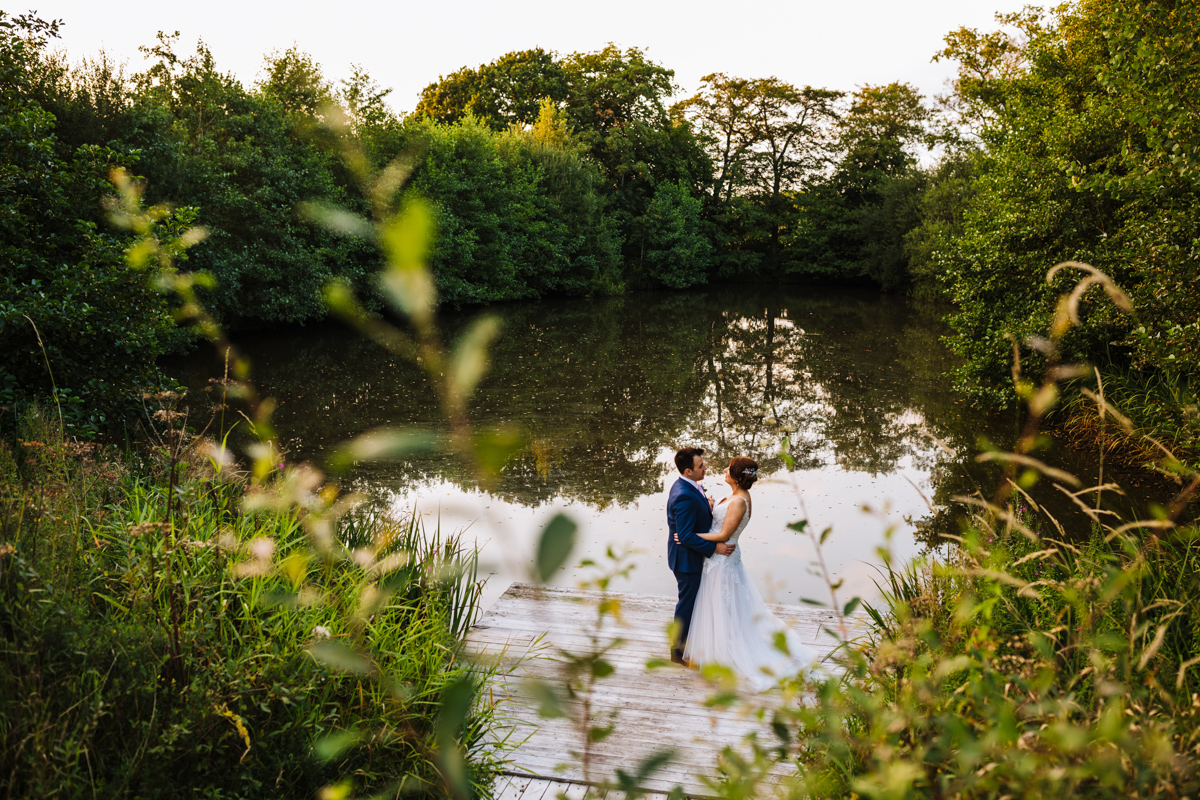 A couple in wedding attire standing close together on a wooden dock by a serene lake surrounded by lush greenery at Styal Lodge