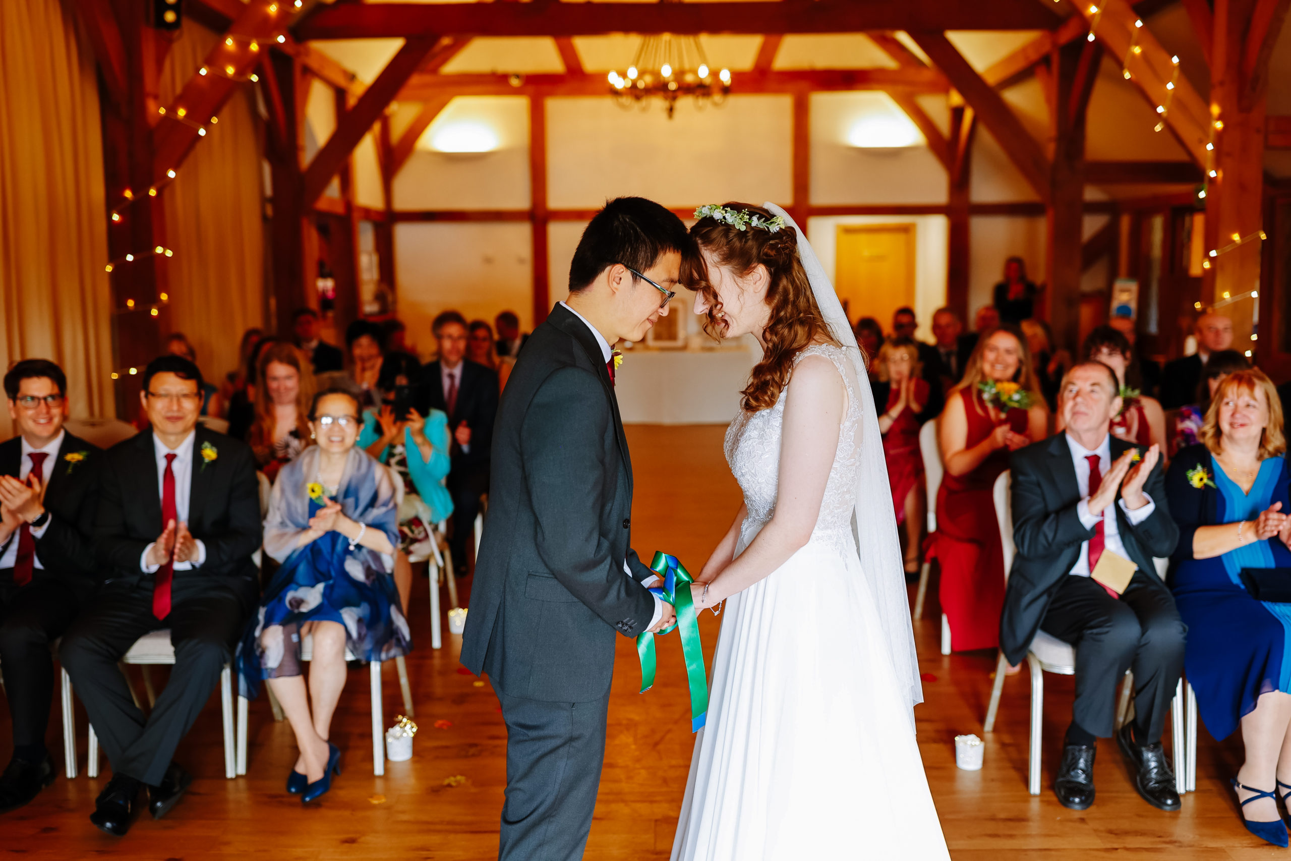 A bride and groom hold hands and touch foreheads in a tender moment during their wedding ceremony, with guests looking on and applauding in a warmly lit venue with wooden beams and string lights.