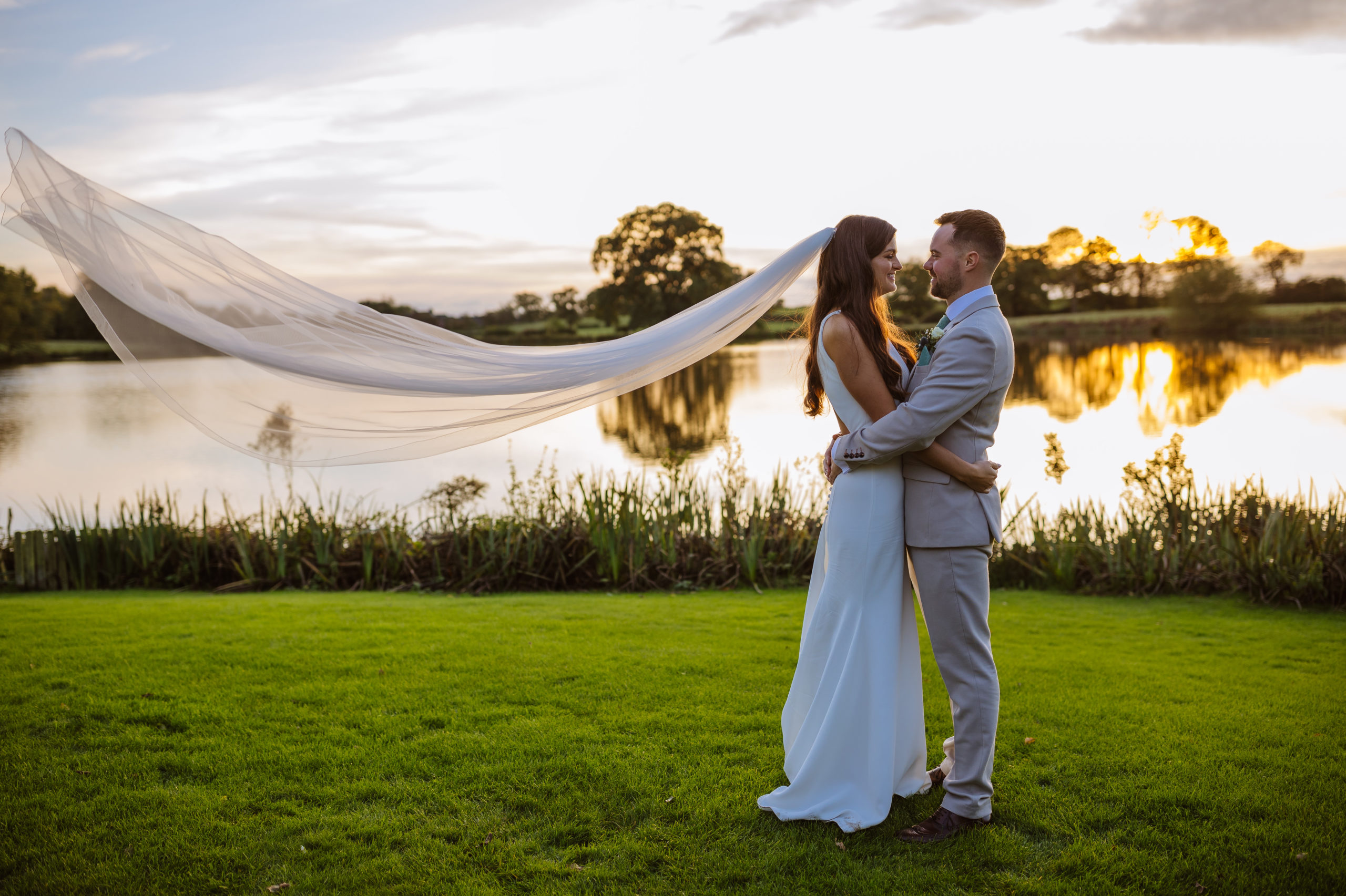 A couple embracing on a grassy lakeside at sunset, with the bride's flowing veil catching the wind.