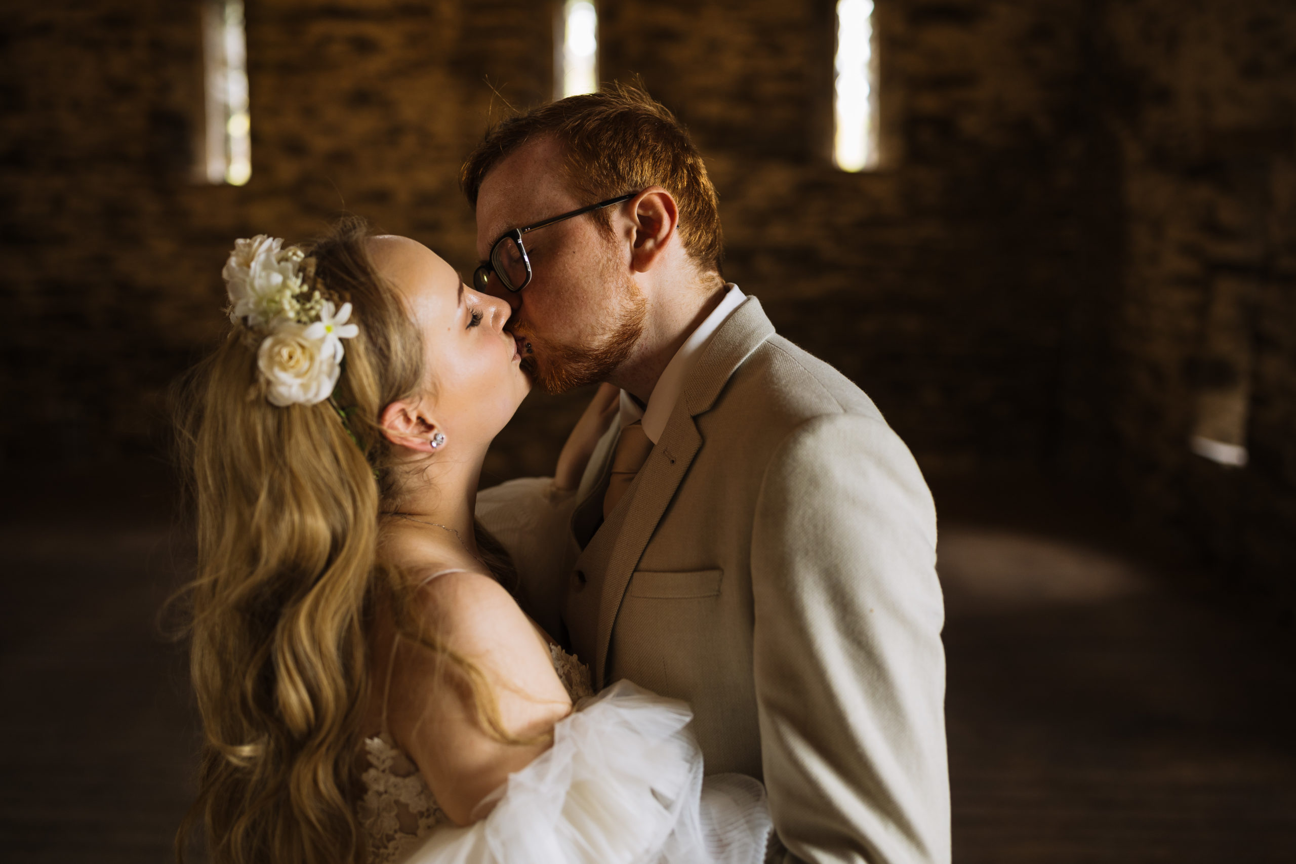 A bride and groom sharing a kiss in a dimly lit room with rustic stone walls, the bride adorned with a floral hairpiece.