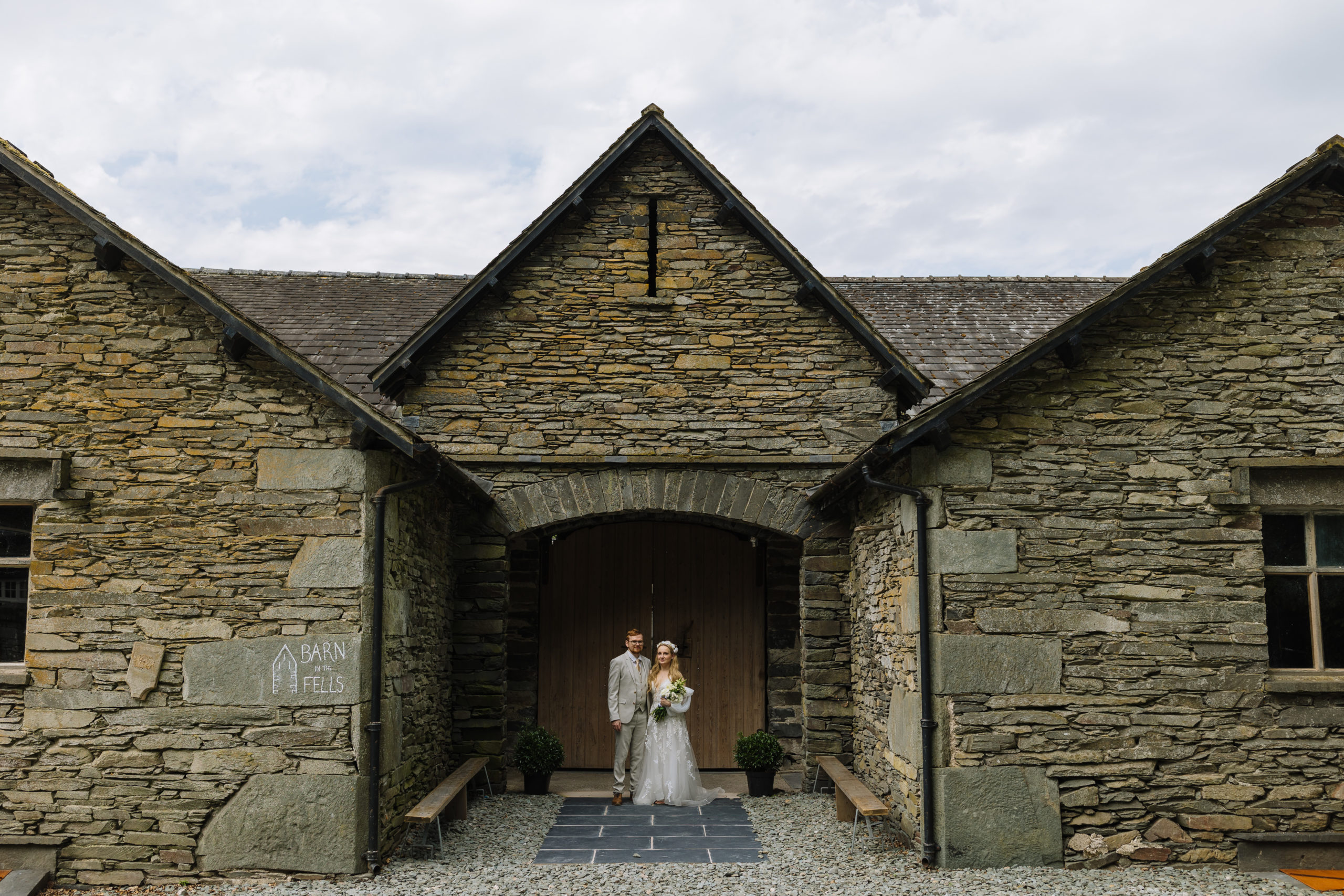 A bride and groom stand holding hands in the entrance of a rustic stone barn with the sign "Barn Fells" on its wall, under a cloudy sky.