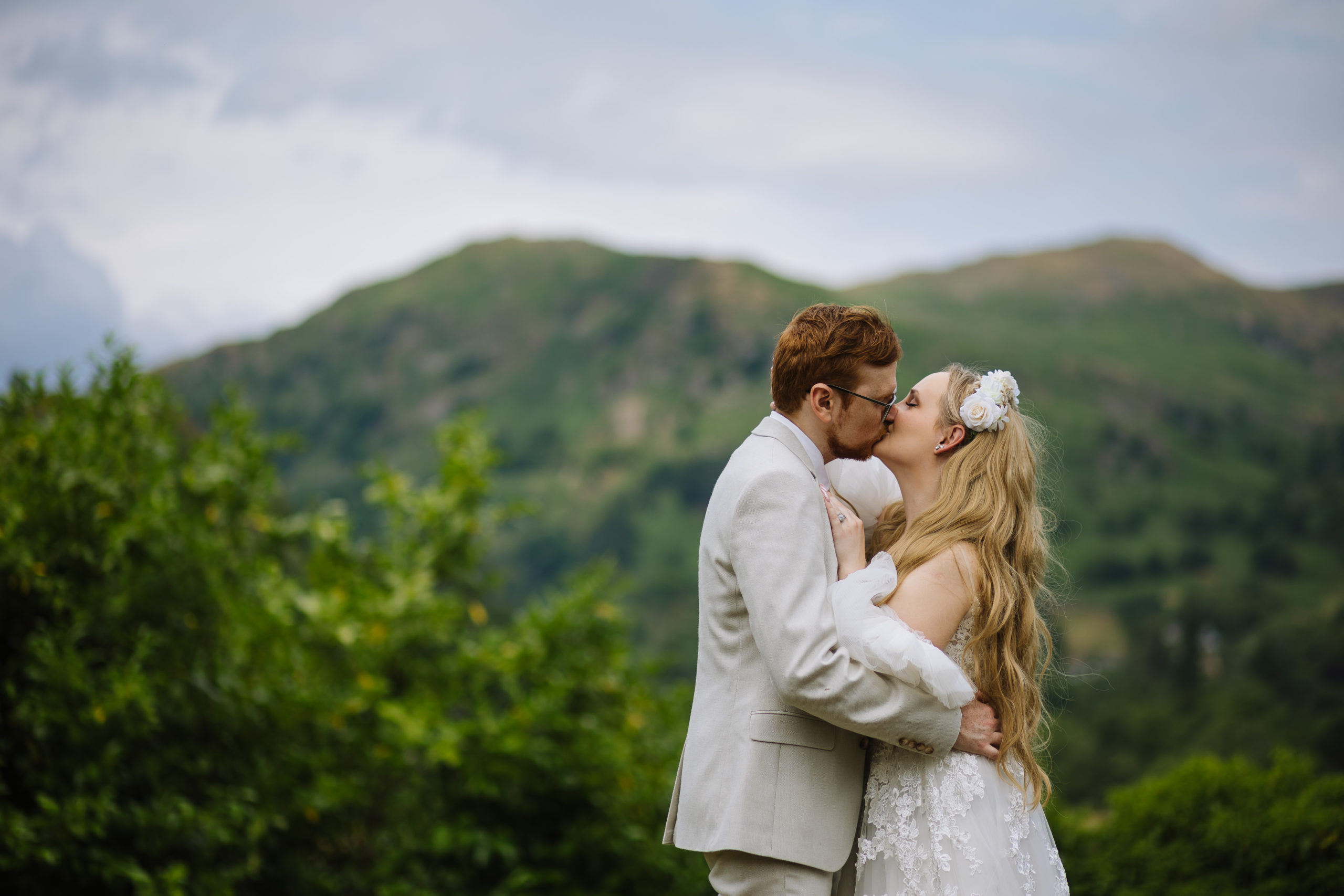 A bride and groom kissing outdoors with a scenic backdrop of green hills and trees.