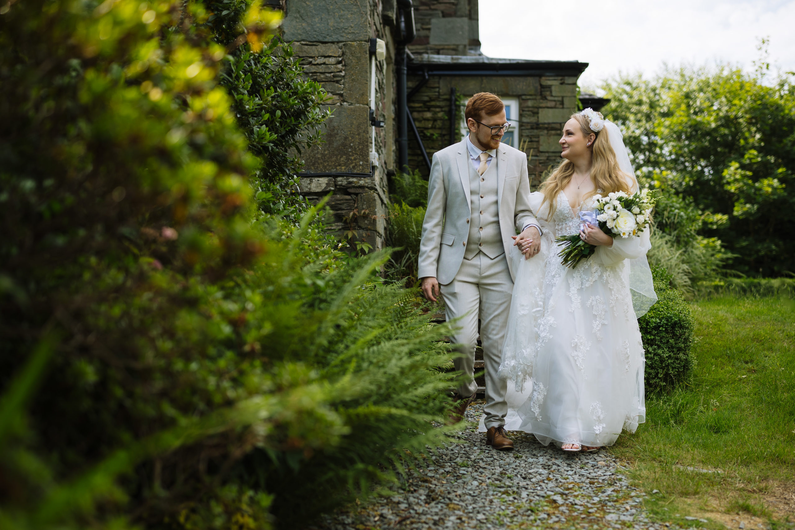 A newlywed couple walking hand in hand along a garden path beside a stone building, with the bride in a white dress holding a bouquet and the groom in a light-colored suit.