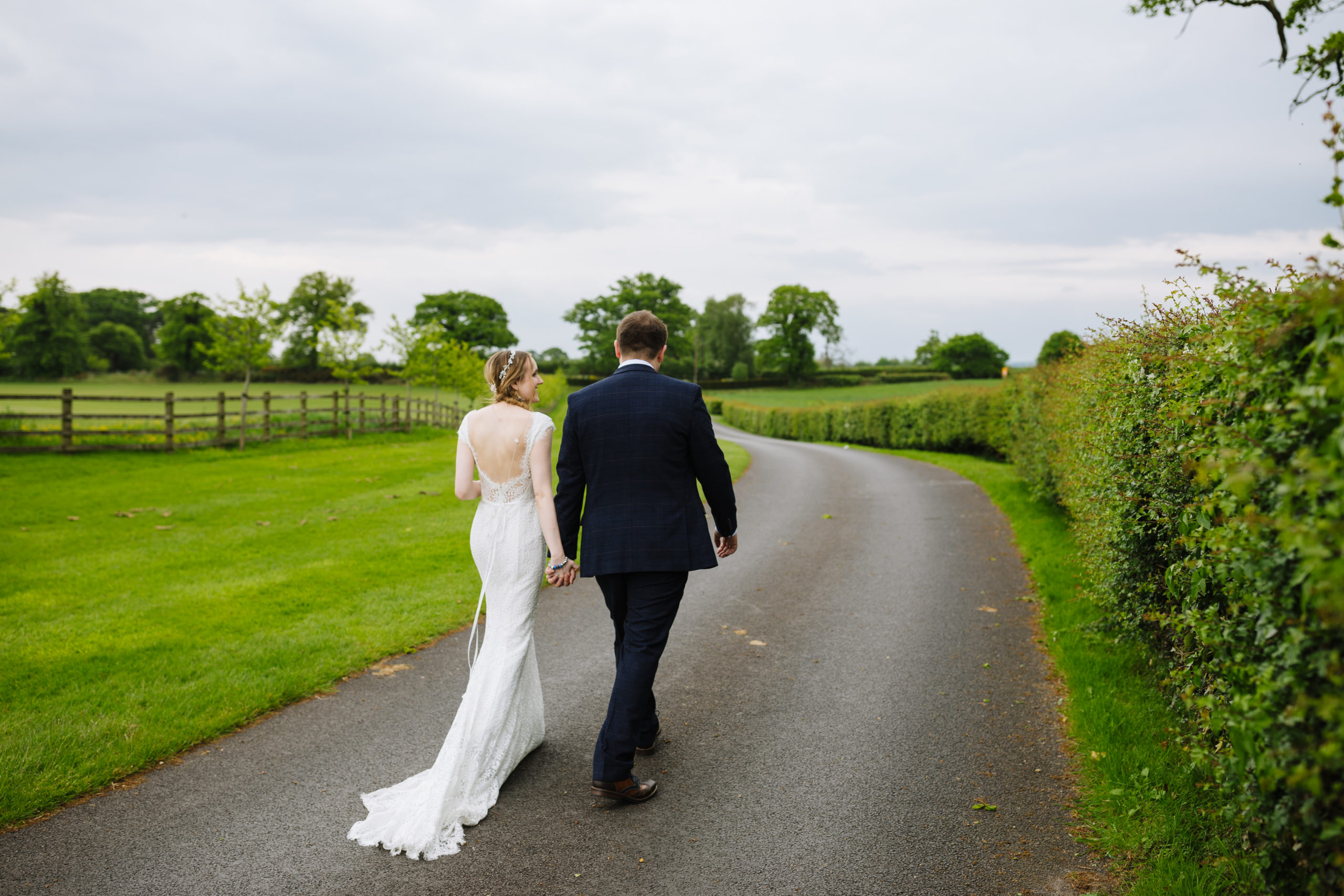 A bride in a white lace gown and a groom in a dark suit walk hand in hand along a rural road, with green fields and hedgerows in the background under a cloudy sky.