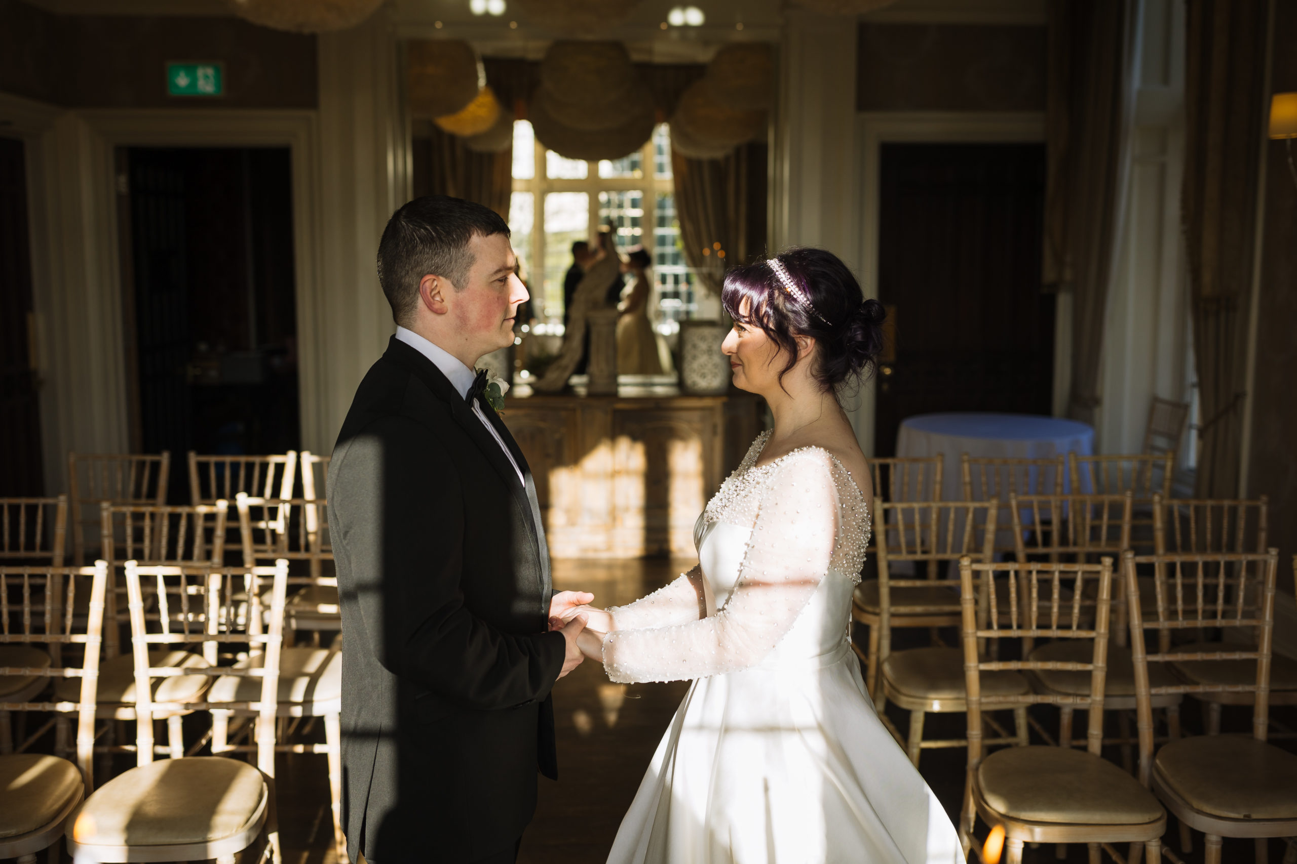 A bride and groom holding hands and gazing at each other in a sunlit room, with rows of empty chairs and elegant decorations in the background.