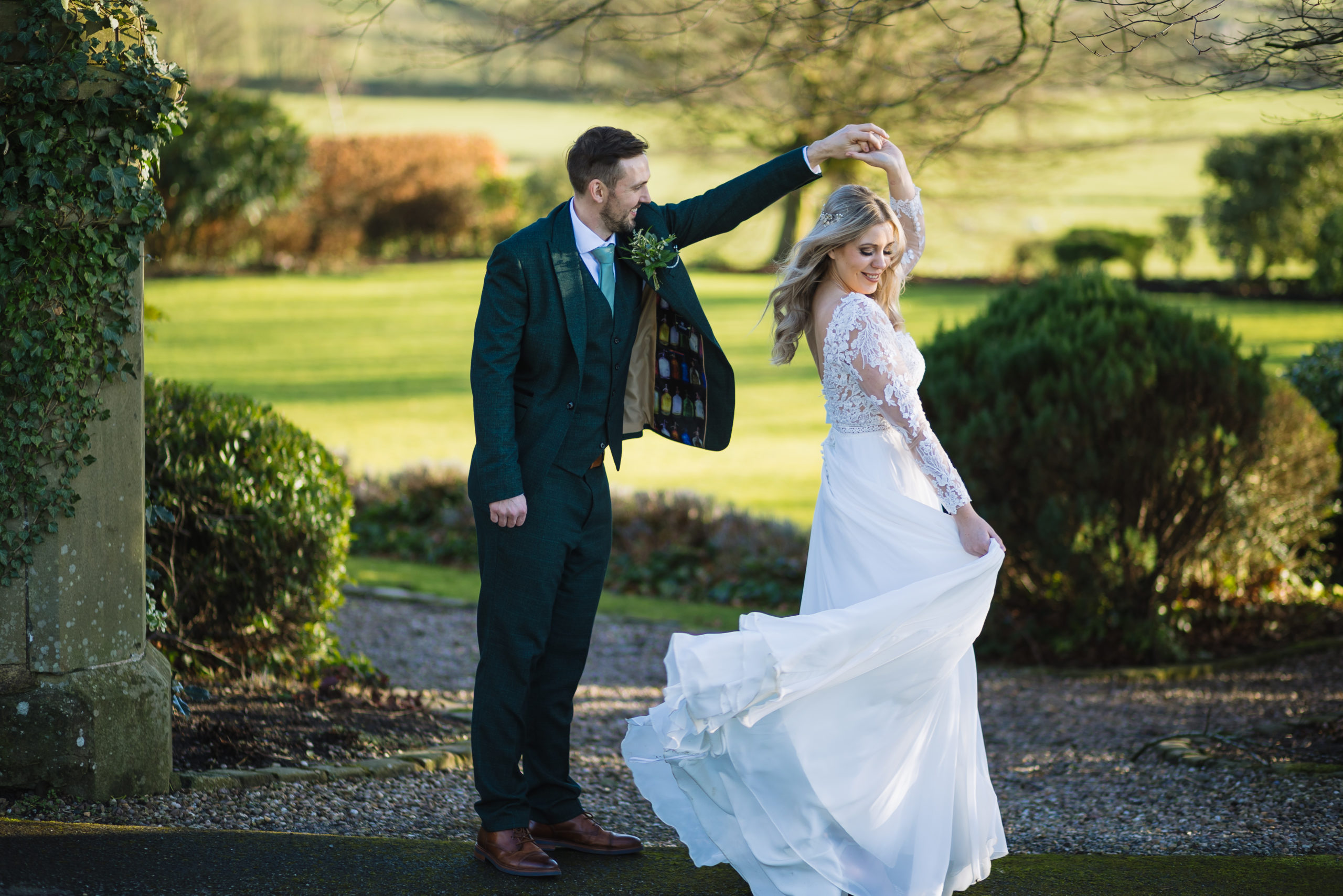 A bride in a long white dress with lace sleeves is dancing with a groom in a dark green suit outdoors, surrounded by greenery and bathed in natural sunlight.