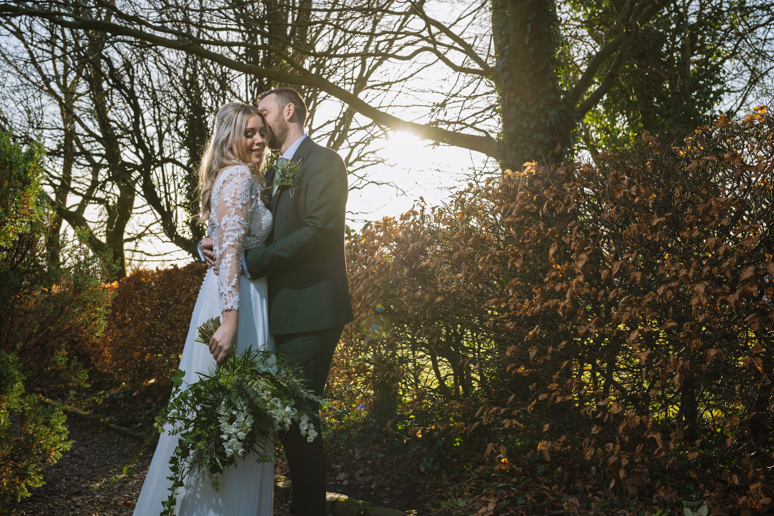 A bride and groom embracing in a sunlit garden with trees and autumn leaves, sharing a joyful moment.