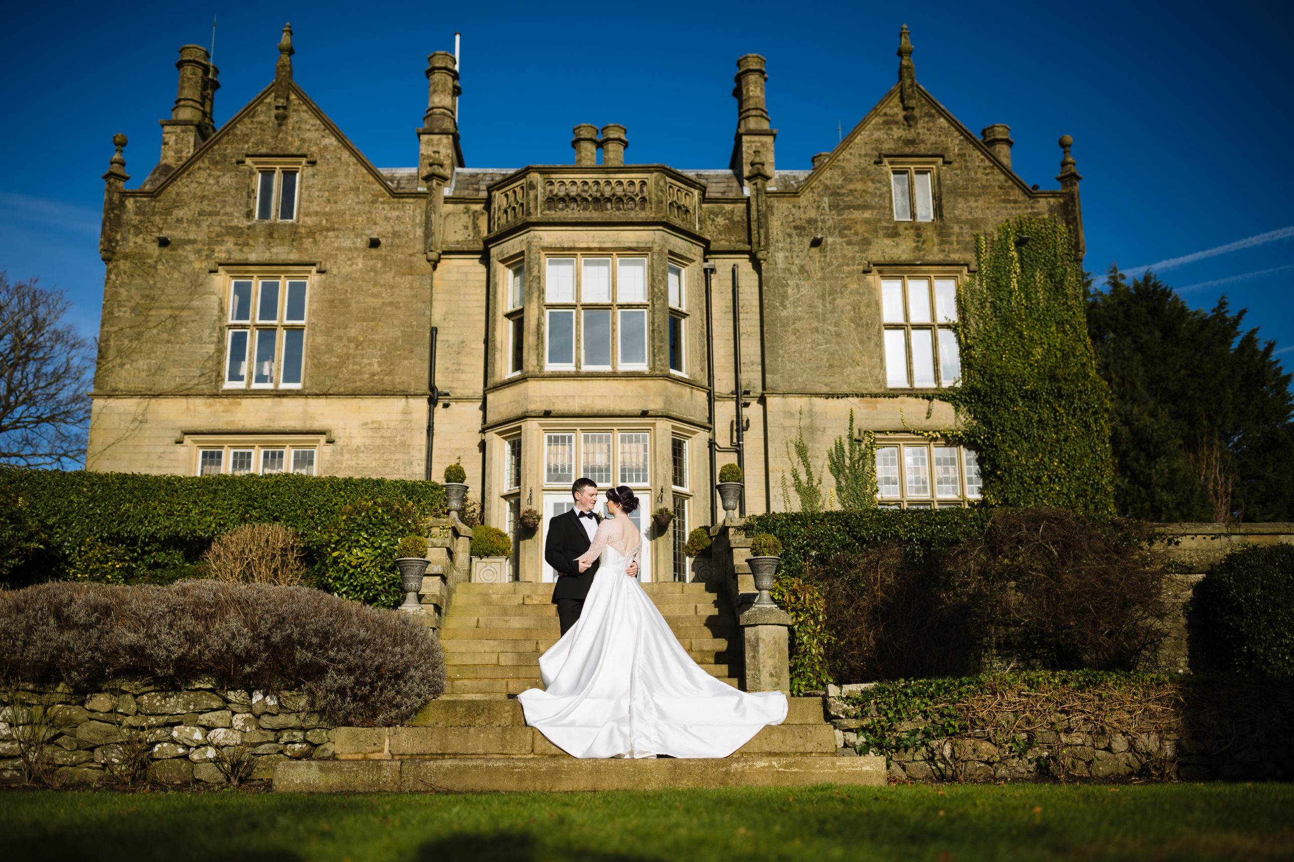 A bride and groom embrace on the steps of an elegant, historic manor on a sunny day, with the bride's dress train flowing down the stairs.