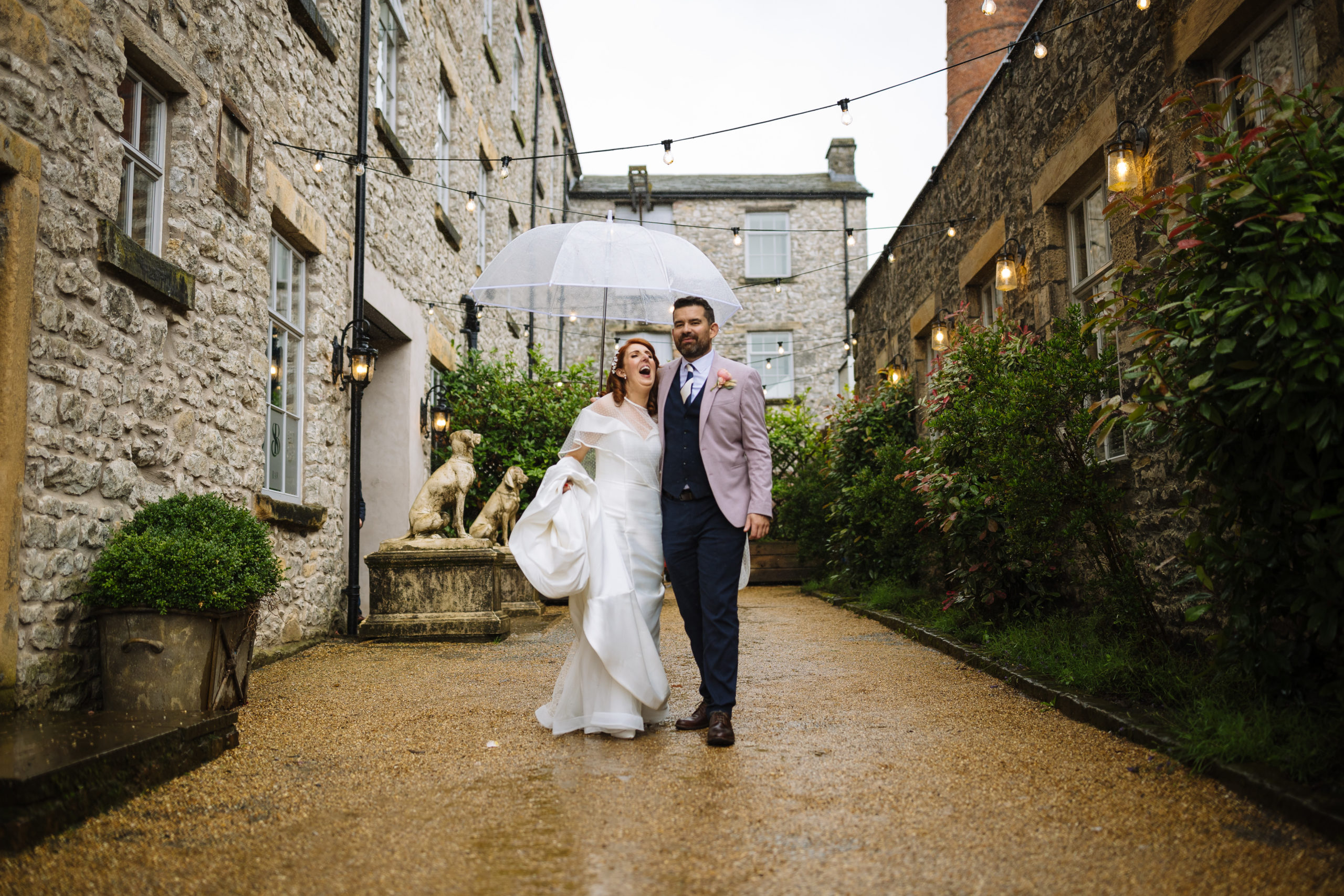 A bride and groom walking hand in hand under a clear umbrella on a rainy day, along a quaint street lined with stone buildings and string lights.