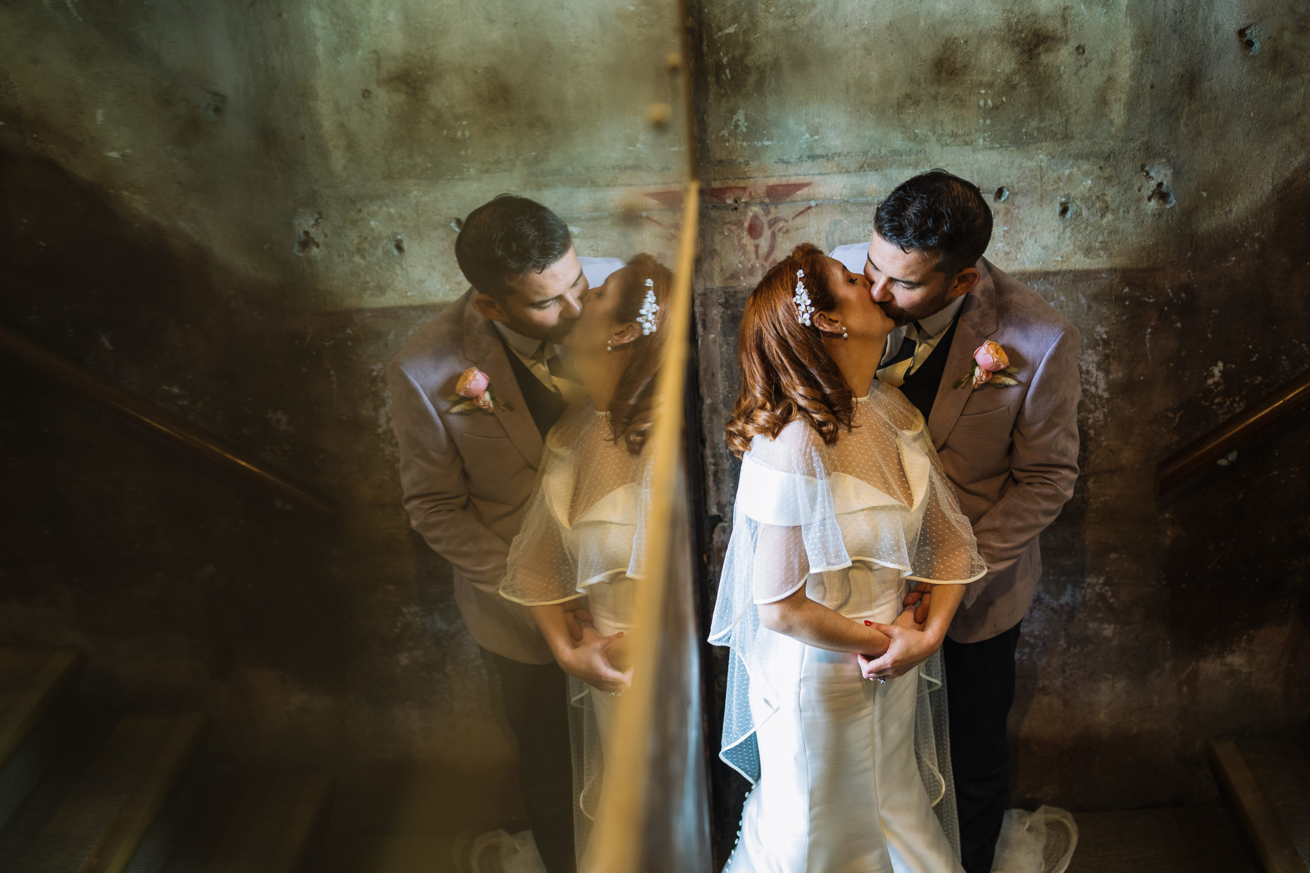 A bride and groom in elegant wedding attire sharing a kiss, reflected in a mirror, creating a double image effect, in a rustic stairwell setting with warm lighting.