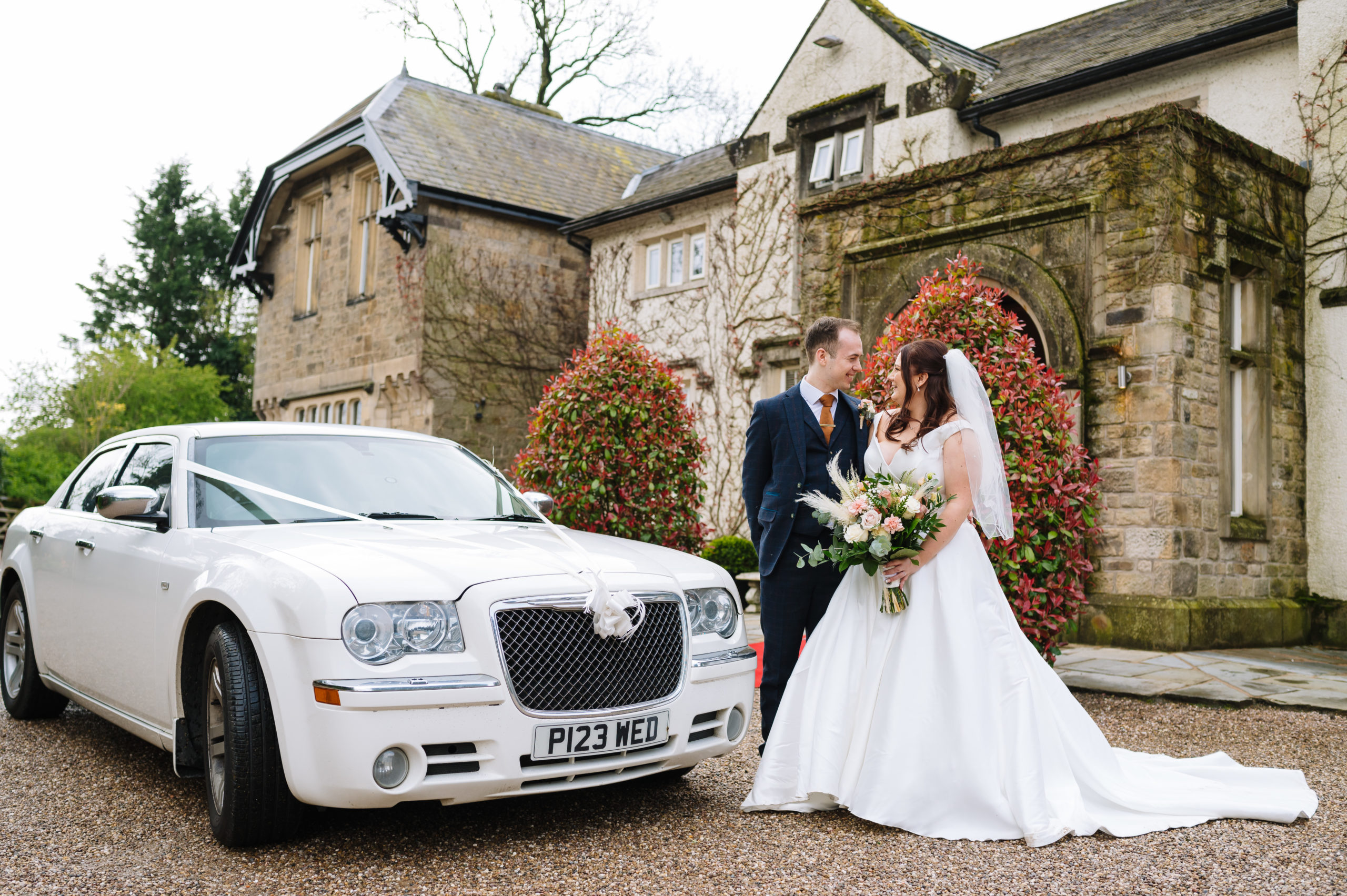 A bride and groom smiling at each other while standing beside a white luxury car, with a historic stone building and red-flowered shrubs in the background.