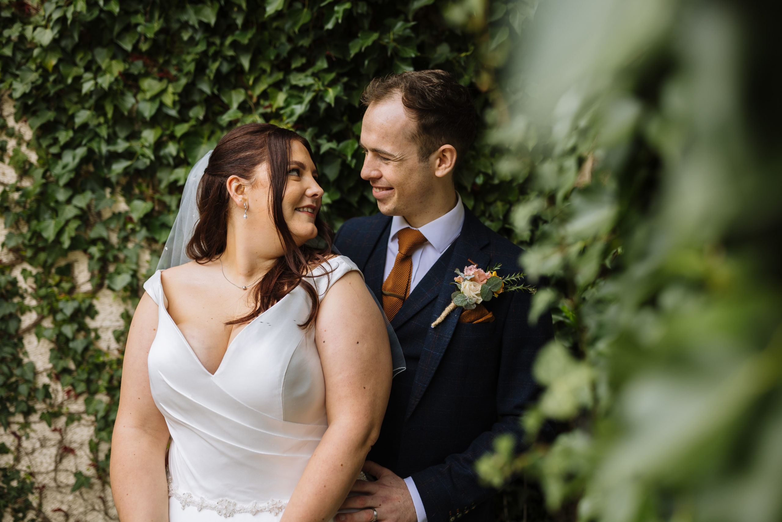 A smiling bride and groom looking at each other, surrounded by greenery, with the bride in a white dress and the groom in a navy suit with an orange tie.