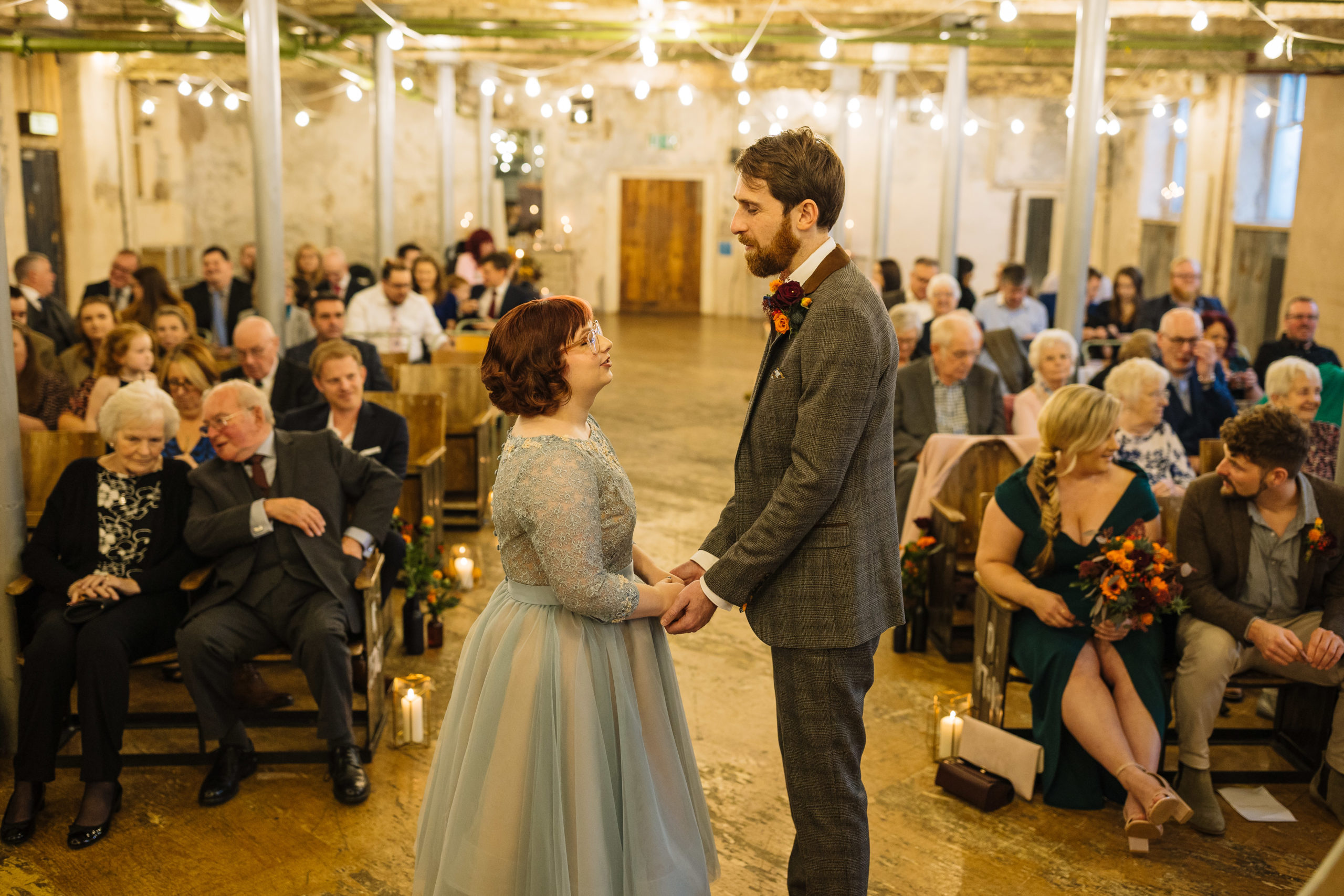 A bride in a blue-gray lace dress and a groom in a gray suit hold hands and look at each other affectionately while standing in the aisle of a warmly lit rustic venue, with guests seated on either side, some looking on with smiles.