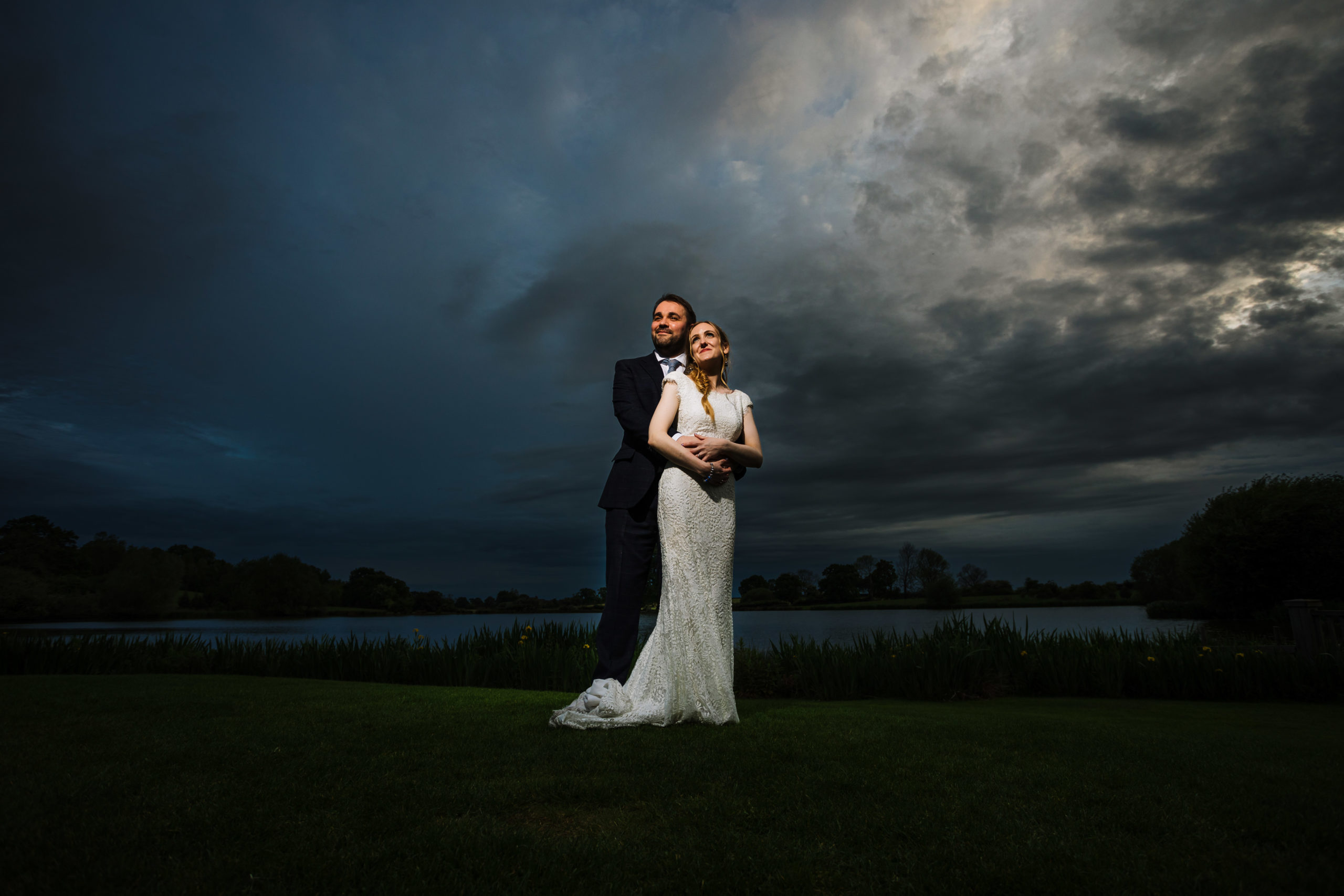 A bride and groom standing together on a grassy field by a lake, with the groom embracing the bride from behind, both gazing into the distance under a dramatic evening sky.