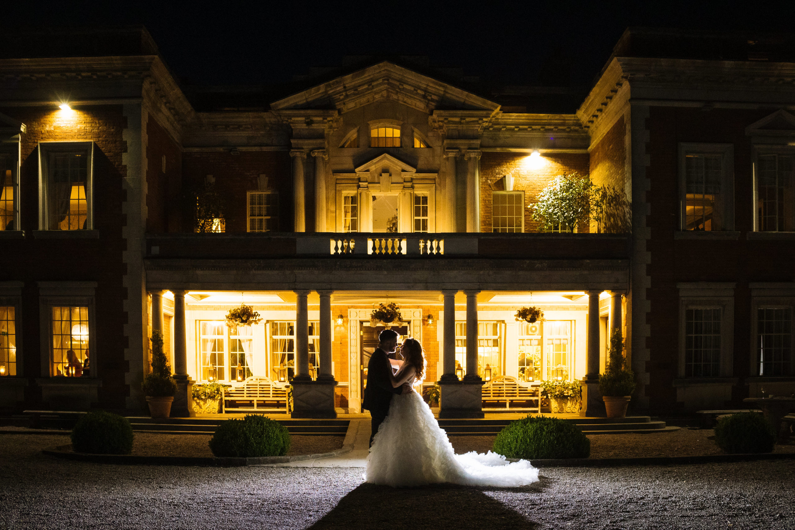 A bride and groom embracing in front of a beautifully lit Eaves Hall during nighttime.