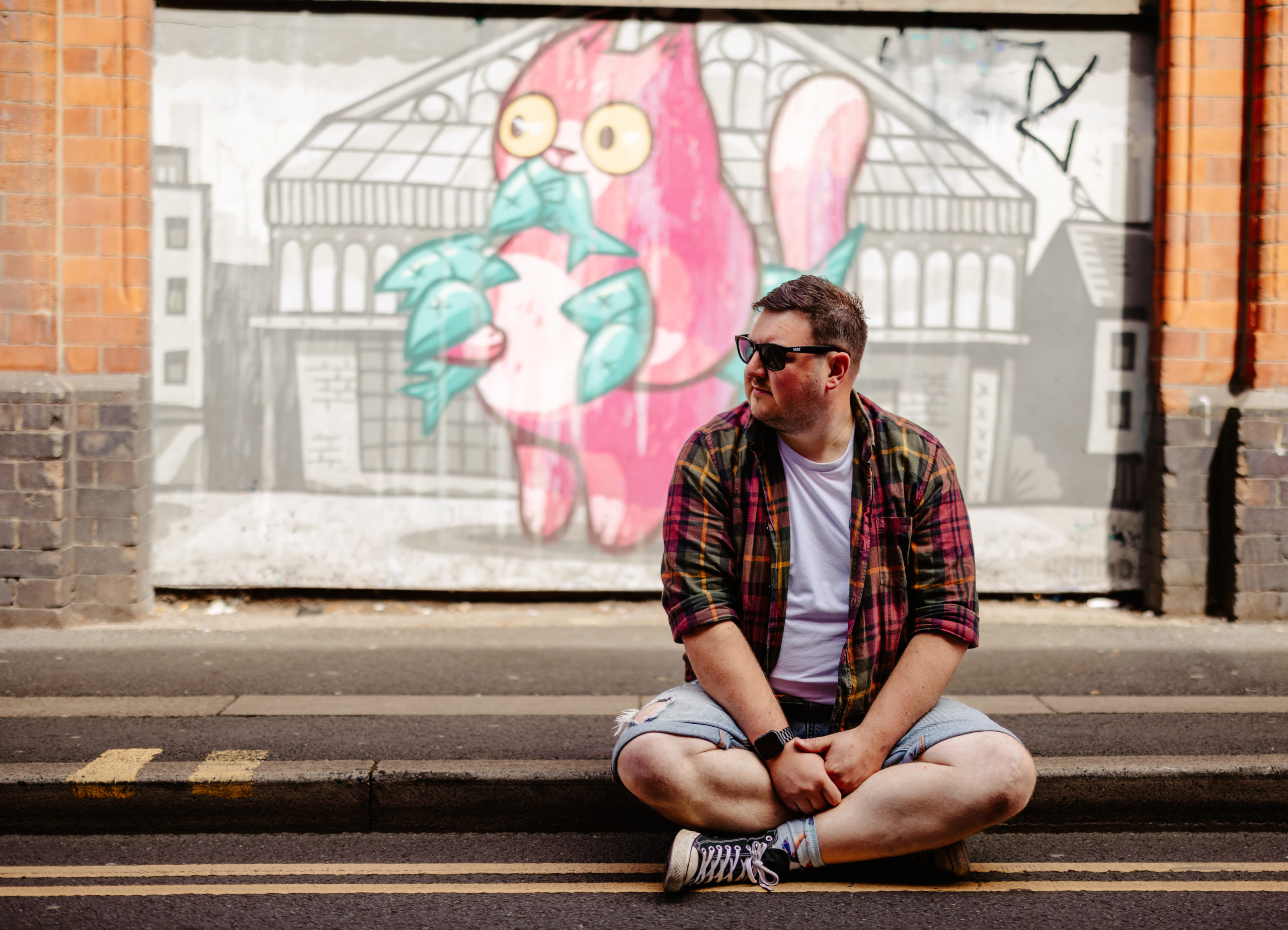 A man in sunglasses and a plaid shirt sits on the street, with a playful pink cat mural in the background.