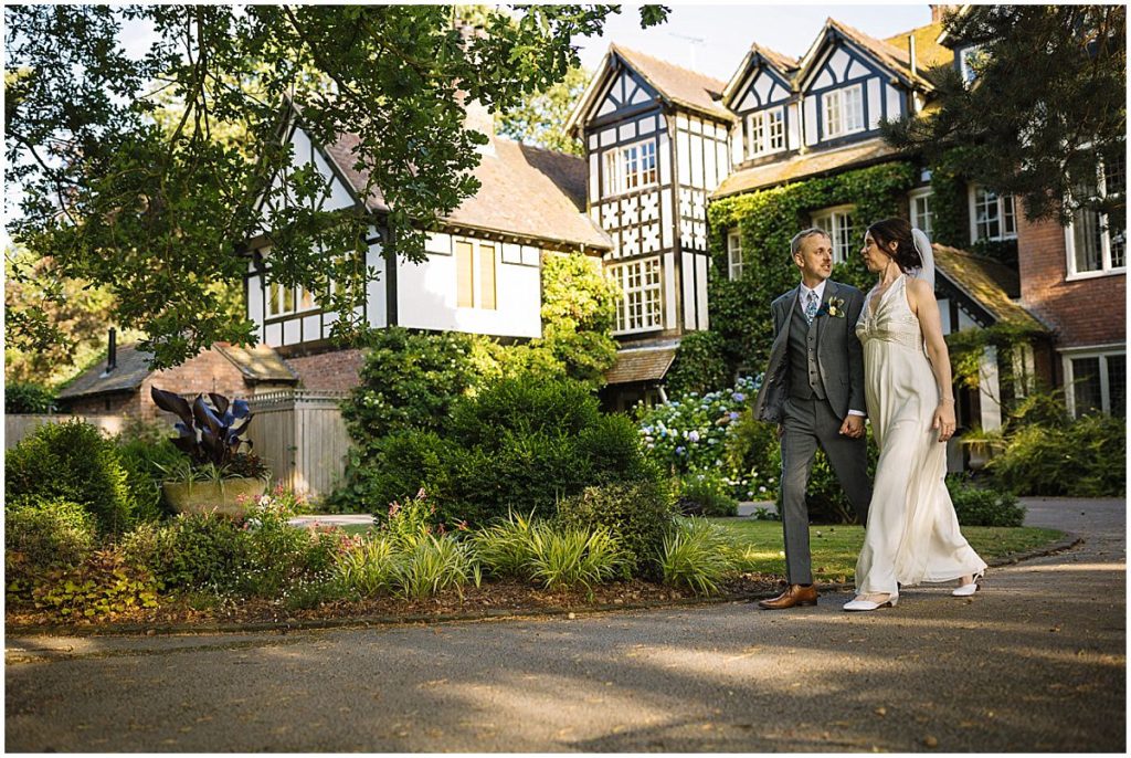 A couple walks hand-in-hand in front of a charming English-style home surrounded by lush greenery, capturing a romantic moment on their wedding day.