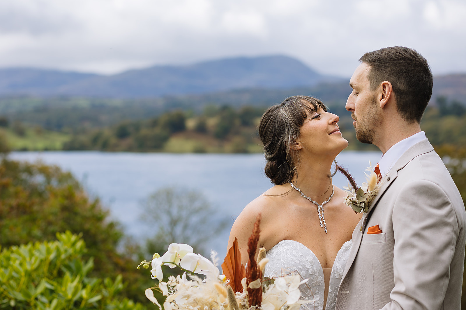 A couple gazes lovingly at each other by a serene lake, surrounded by lush greenery and distant mountains, capturing a beautiful wedding moment.