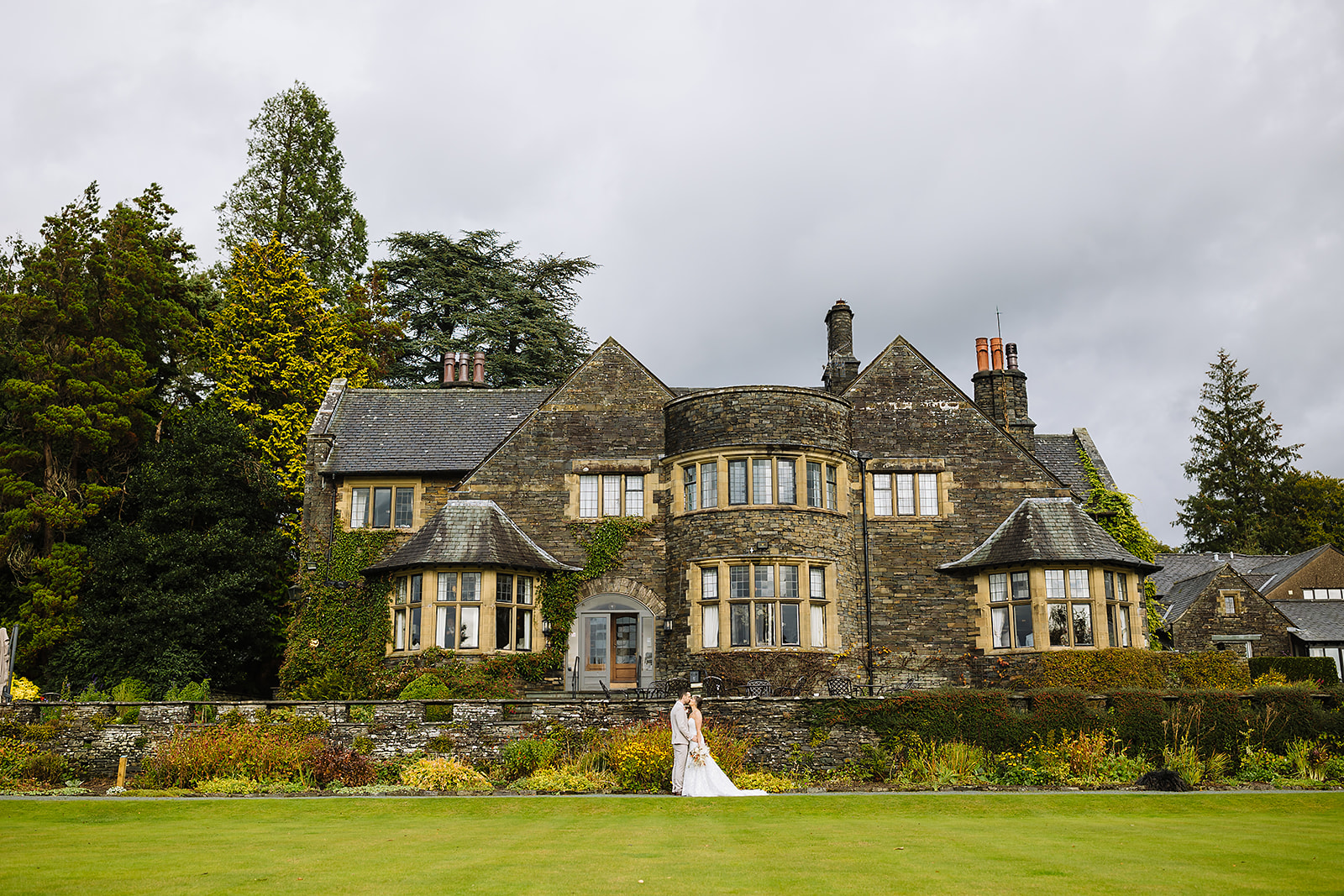 A bride standing in front of a grand stone mansion surrounded by lush gardens and tall trees under a cloudy sky in the Lake District